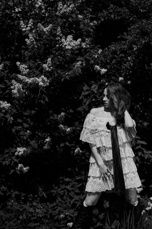 Beautiful young woman with long curly hair in white lace dress standing in blooming lilac garden.の写真素材