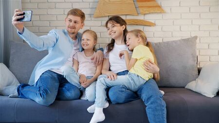Happy parents and twins smiling and taking selfie while sitting on comfortable sofa at home togetherの写真素材