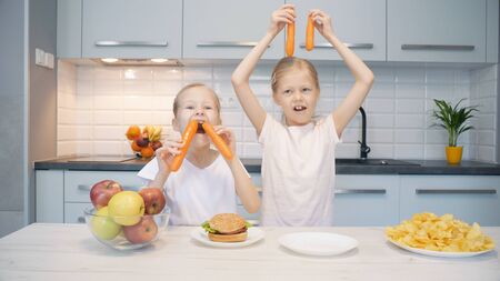 Two cute little girls grimacing and playing with fresh carrots over table with apples and junk food at homeの写真素材
