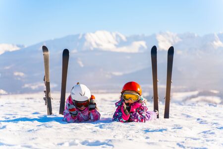 Children in helmets and goggles lying on snowy terrain with skis near on background of mountainsの写真素材