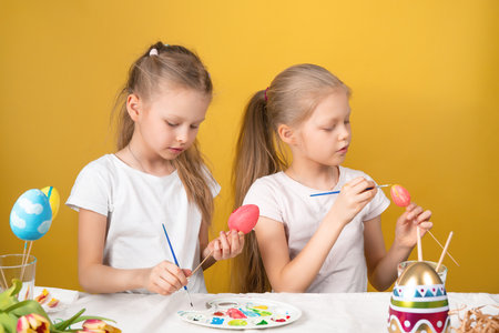 Cute little sisters with colorful eggs sitting at table with palette and Easter symbols against yellow backgroundの写真素材