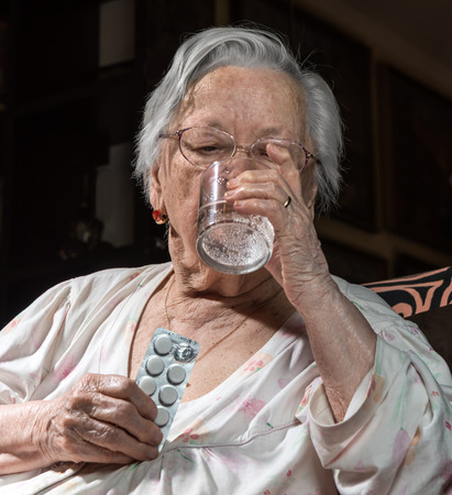 Old sick woman with pills and glass of water at homeの写真素材