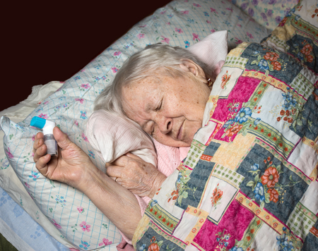 Old sick woman with asthma inhaler lying in bed at homeの写真素材