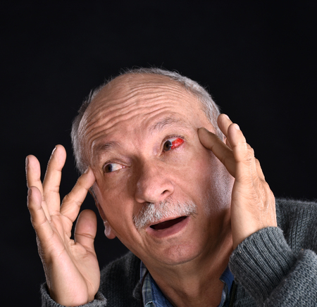 Senior man with irritated red bloodshot eye suffering from pain on a grey background