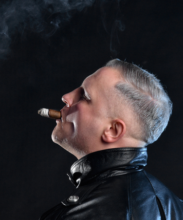Portrait of handsome man with a cigar in leather jacket posing  in a dark roomの写真素材