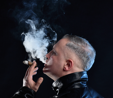 Portrait of handsome man with a cigar in leather jacket posing  in a dark roomの写真素材