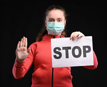 Pandemic coronavirus concept. Teen girl in protective sterile medical mask on her face showing stop sign over black backgroundの写真素材