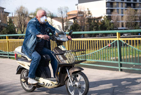 UZHHOROD, UKRAINE - APRIL 10, 2020: People in medical masks on the street rush about their business during the coronavirus epidemic. Elderly man on a motorcycleのeditorial素材