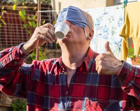 Senior man in protective mask drinking coffee in the garden at homeの写真素材
