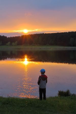The boy stands on the shore of the lake, looking at the setting sun.の写真素材