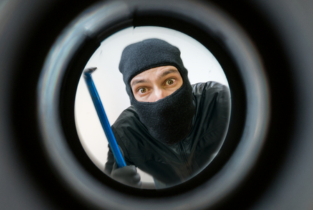 View through pipehole. Thief or burglar masked with balaclava is holding crowbar behind the door.の写真素材