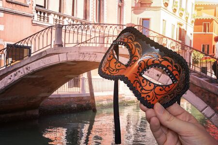 Hand is holding Venetian carnival mask at famous traditional venetian festival in Italy. Bridge over canal  in background.の写真素材
