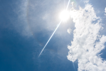 Sun shine with rainbow ring radiation behind white clouds in blue skyの写真素材