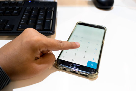 Finger of man who is using the calculator application in mobile phone on the white deskの写真素材