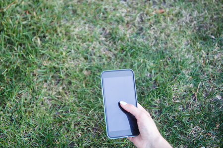 The phone in the girls hand against the background of the green grass. The girl is writing a message in the park.の写真素材