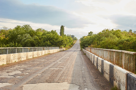 Damaged asphalt pavement road with potholes caused by freeze and thaw cycle during winter. Bridge over the river. Damaged asphalt in Ukraine.の写真素材