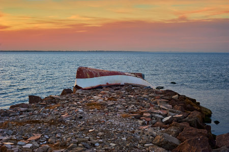 Old abandoned damaged fishing boat laying on the stone pier. Beautiful sunset on the lake on Summer eveningの写真素材