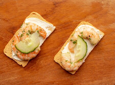 Two crispbreads, cream-cheese, prawns and cucumber on a wooden boardの写真素材