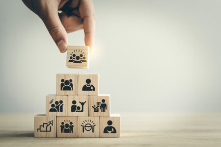 A hand places a wooden block with a team icon onto a pyramid of business concept blocks. This symbolizes strategic planning, leadership, and teamwork for organizational growth. It represents building success and fostering innovationの素材