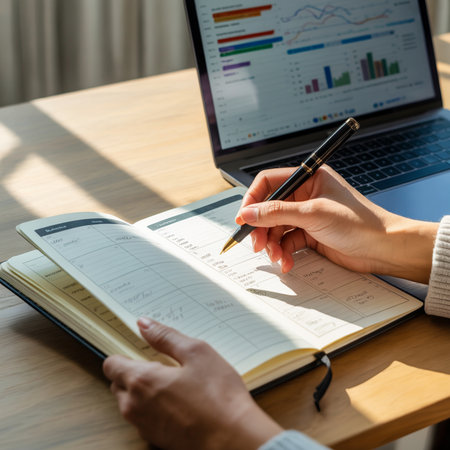 Close up of hands writing in an open planner next to a laptop displaying colorful charts and graphs on a wooden desk with sunlight casting shadows.の素材