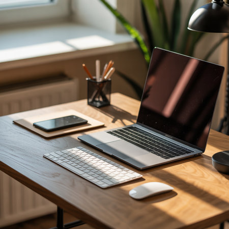 A wooden desk holds an open laptop, keyboard, mouse, and smartphone. Pencils in a holder and a green plant are visible. Sunlight casts shadows across the surface.の素材