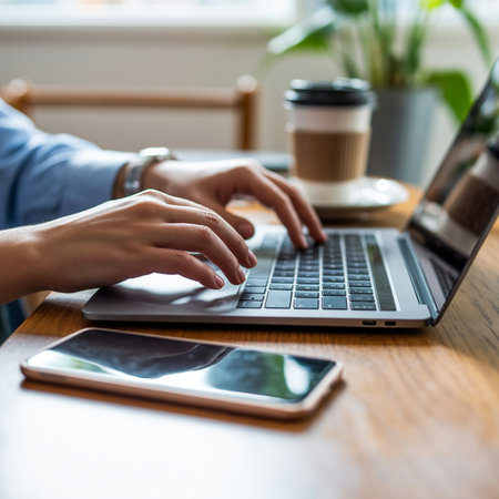 Hands typing on a laptop keyboard on a wooden table with a smartphone and a coffee cup nearby. Natural light illuminates the scene.の素材
