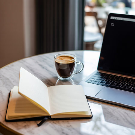 Open notebook with blank lined pages and a glass mug of dark coffee sit on a reflective marble table next to a silver laptop.の素材
