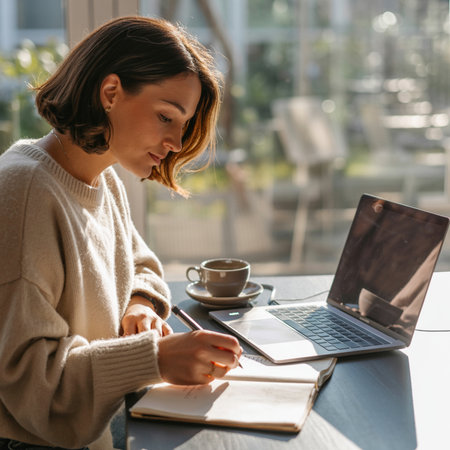 Woman with short brown hair in a cream sweater writes in an open notebook with a pen. A laptop and coffee cup sit on a dark table in front of a sunlit window.の素材