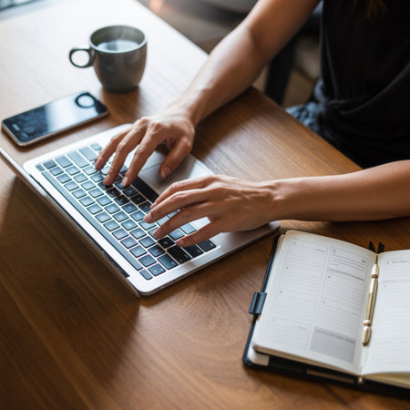 Hands typing on a silver laptop keyboard on a warm brown wooden desk. A grey mug of hot coffee and a smartphone sit nearby, along with an open notebook and pen.の素材
