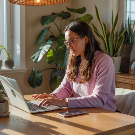 Woman with glasses and pink sweater types on a silver laptop at a wooden desk. Sunlight streams through a window illuminating large green houseplants.の素材