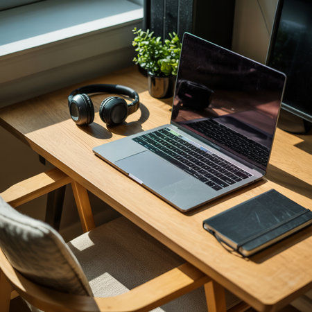 A laptop is open on a light wood desk next to black headphones and a black notebook. Sunlight streams through a window, casting shadows on the desk and a chair.の素材