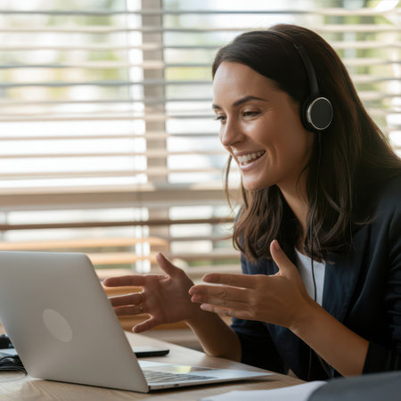 Woman with dark hair wearing headphones and a dark suit smiles while gesturing with her hands during a video call on a silver laptop.の素材