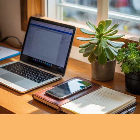 A laptop, smartphone, and notebooks rest on a wooden desk bathed in sunlight from a nearby window. Two potted succulents add a touch of green to the scene.の素材