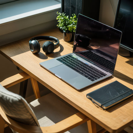 A wooden desk with a laptop, black headphones, a dark notebook, and a small green plant sits near a window casting shadows.の素材
