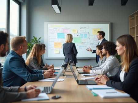 A group of business professionals sit around a wooden table in an office setting, engaged in a meeting. Laptops and papers are scattered across the table. A whiteboard with colorful sticky......の素材