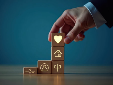 A hand in a business suit sleeve places a wooden block with a heart symbol on of a stack of wooden blocks with various including a house, profile, and graph, on a table against a blue background......の素材