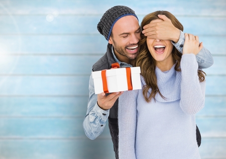 Happy man holding a gift box and covering eyes of woman against wooden backgroundの写真素材