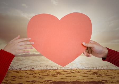 Hand of couple holding red heart on beach against digitally generated backgroundの写真素材