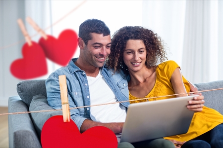 Composite image of hanging red hearts and couple using a laptop on sofaの写真素材