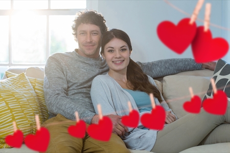 Composite image of red heart and happy couple embracing on sofa in living roomの写真素材