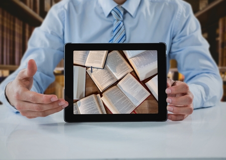 Digital composite of Business man at table with tablet showing open books against blurry bookshelvesの写真素材
