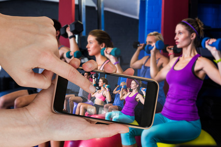 Friends holding dumbbells while sitting on fitness ball  against hands touching smart phone