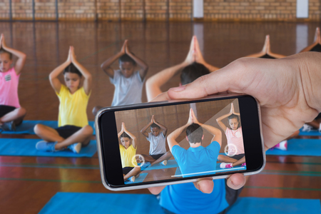 Hand holding mobile phone against white background against school kids and teacher meditating during yoga classの写真素材