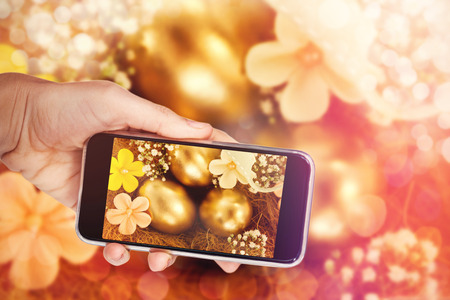 Hand holding mobile phone against white background against golden easter eggs with flowers in nestの写真素材