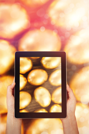 Close-up of hands holding digital tablet against close-up of golden easter eggsの写真素材