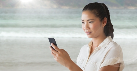 Digital composite of Woman looking at phone against blurry beach and flareの写真素材