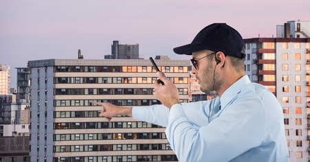 Digital composite of Security guard holding walkie talkie pointing against buildings