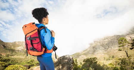 Digital composite of Hipster carrying backpack with camera looking at mountain while standing on cliff against skyの写真素材
