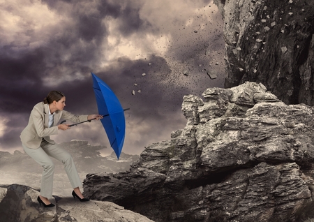Digital composite of Digital image of rocks falling on businesswoman with blue umbrella standing against skyの写真素材