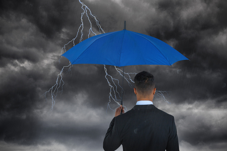 Rear view of businessman carrying blue umbrella  against stormy skyの写真素材